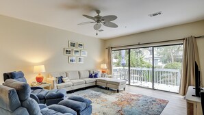 Sala de estar con sofá, sillas, ventilador de techo y un amplio ventanal corredera al exterior.
