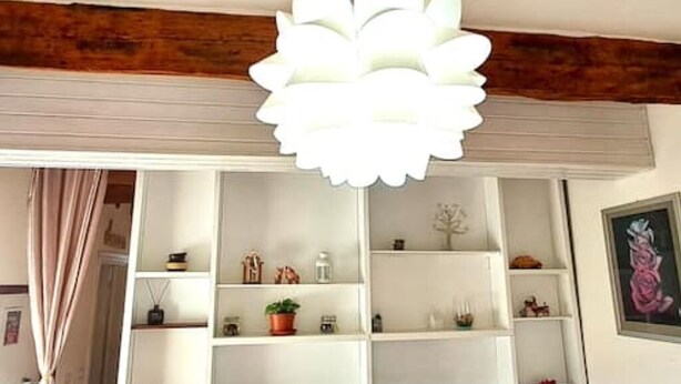 Una stanza con soffitto bianco, travi in legno, una scaffalatura bianca con diversi oggetti e una lampada a forma di fiore bianca.