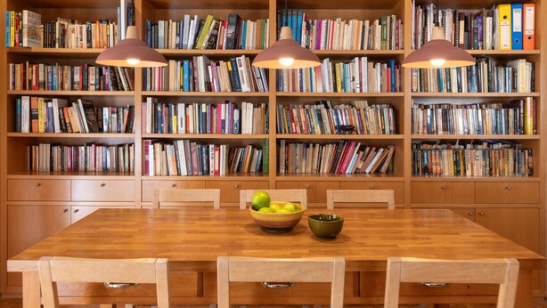 Ein Bücherregal aus Holz voller Bücher, ein Holztisch mit Stühlen und eine Obstschale.