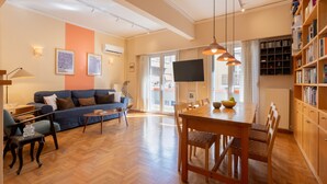 Ein Wohnzimmer mit einer Couch, einem Couchtisch und einem Esstisch mit Stühlen. Ein Bücherregal, ein Flachbildfernseher und ein Fenster mit Vorhängen sind ebenfalls vorhanden.