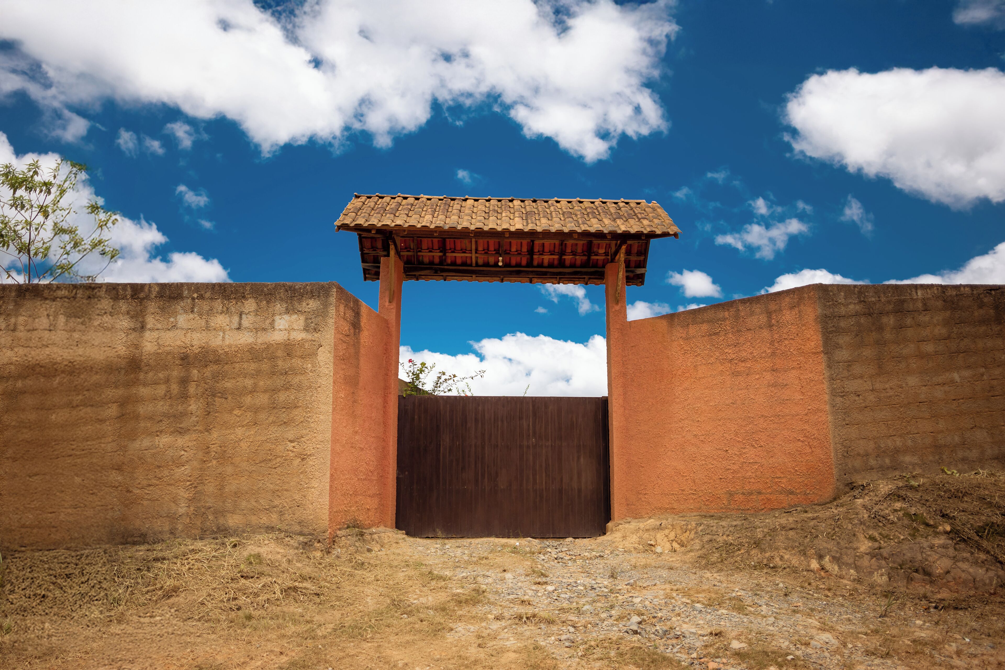 Una porta rústica amb teulada de fusta, envoltada de murs de rajola, sota un cel blau i clar.