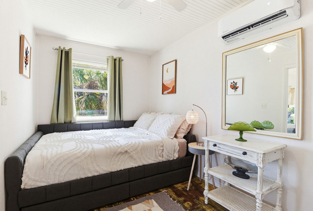 Ein Schlafzimmer mit einem Bett, einem Nachttisch, einem Spiegel und einem Fenster mit Vorhängen.