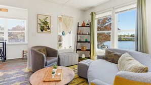 Ett vardagsrum med en soffa, fåtölj och pall, ett träbord, en bokhylla och utsikt över en sjö eller ett annat vattendrag.