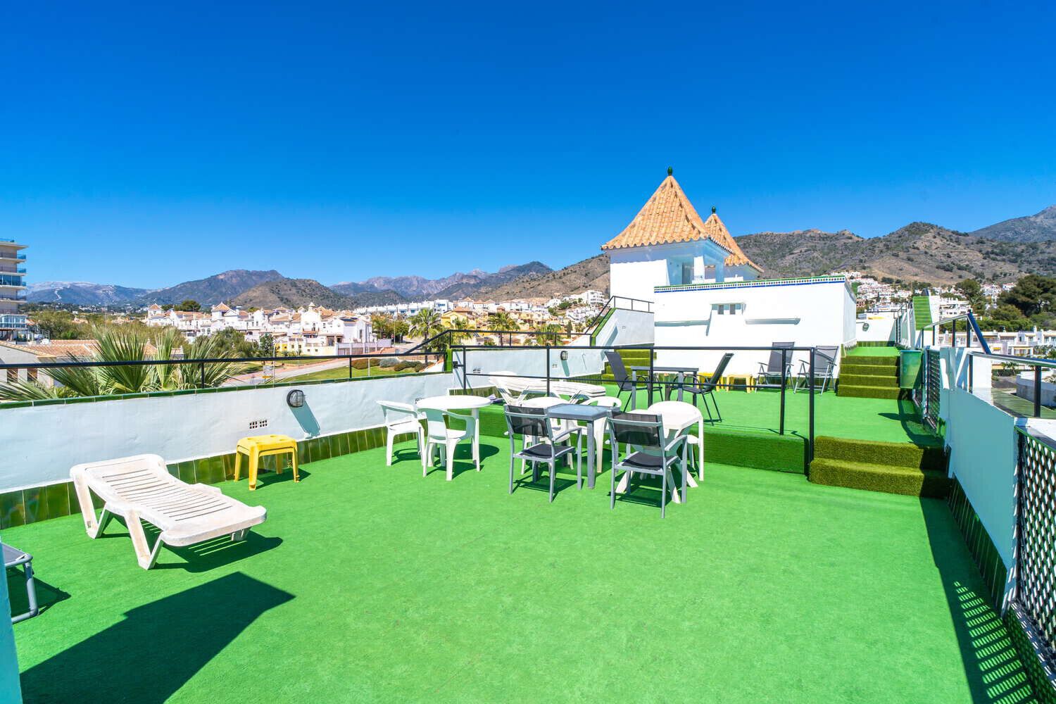 A rooftop terrace with green artificial turf, white lounge chairs, and tables, overlooking a coastal town and mountains.