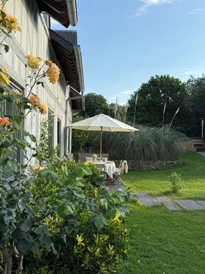 Une terrasse avec une table et des chaises, un grand parasol et un jardin où poussent différentes espèces de plantes.