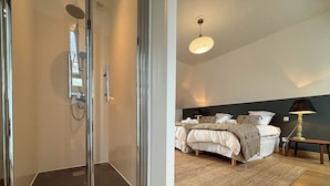 Une chambre d’hôtel moderne dotée d’une douche en verre, d’un lit avec une tête de lit sombre et d’un tapis tissé.