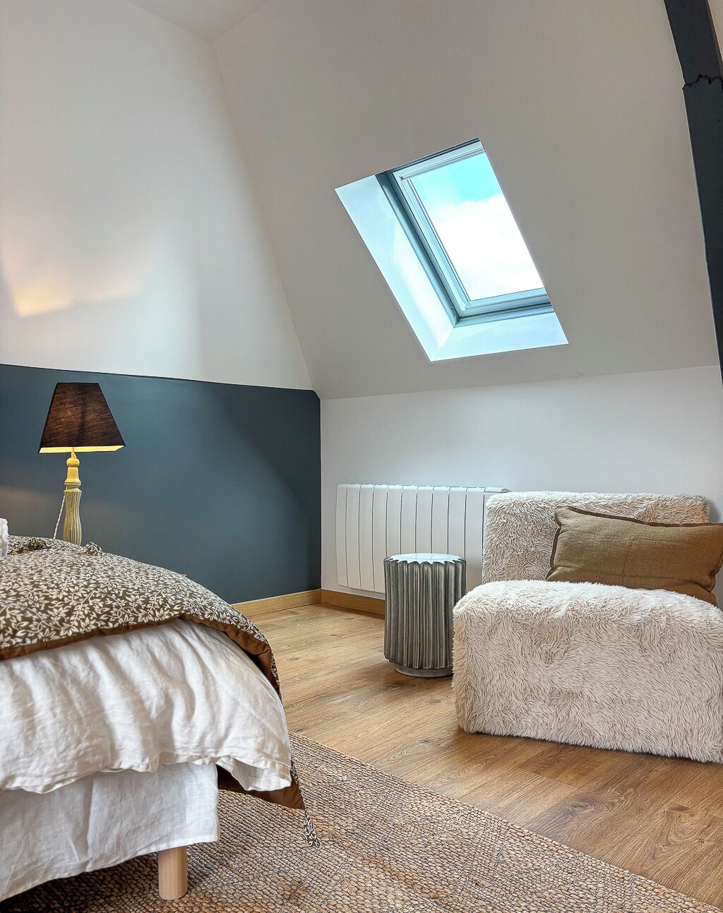 A bedroom with a skylight, a bed, a bedside table with a lamp, a radiator, and a fluffy ottoman.