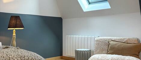 A bedroom with a skylight, a bed, a bedside table with a lamp, a radiator, and a fluffy ottoman.