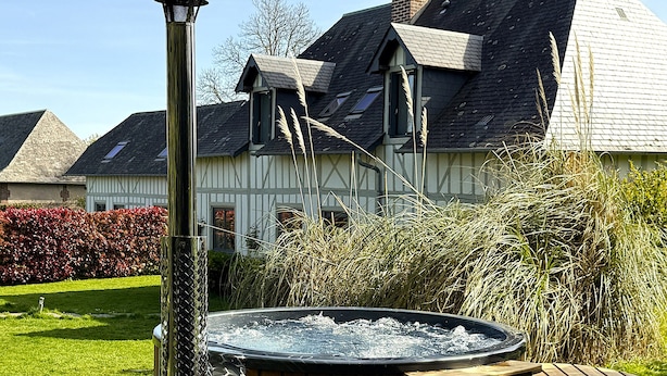 Un bain à remous avec une structure extérieure en bois, une cheminée en métal et, au fond, une maison avec un toit en pente.