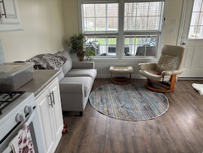 A modern kitchen with a built-in oven, a gray sofa, a beige leather chair, and a large window with a view of the outside.