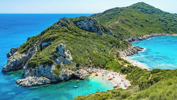 Paisatge costaner amb una illa rocosa, aigües turqueses cristal·lines i una platja amb gent.