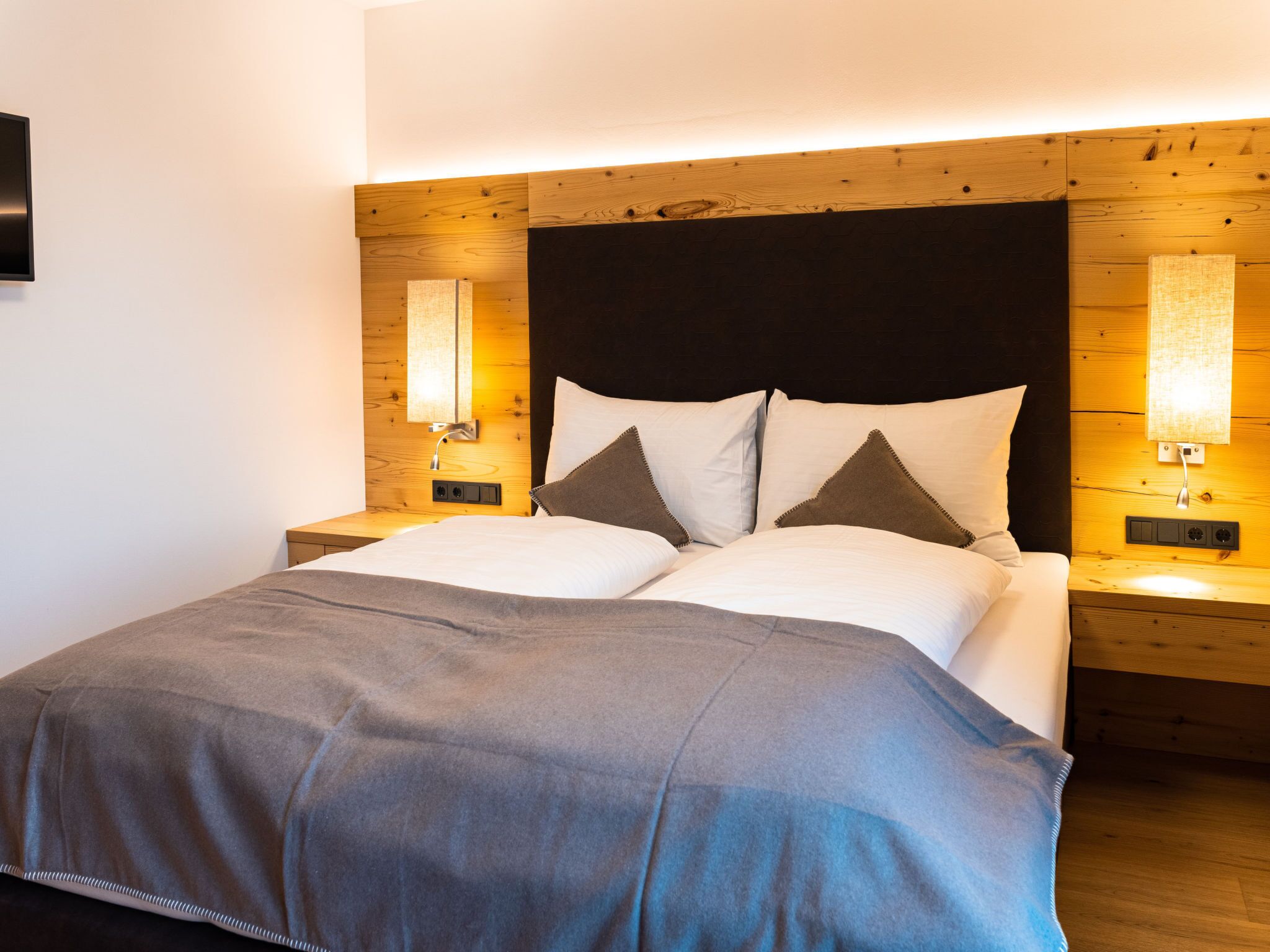 Een netjes opgemaakt bed met een grijze deken, twee nachtkastjes met lampen en een houten hoofdbord.