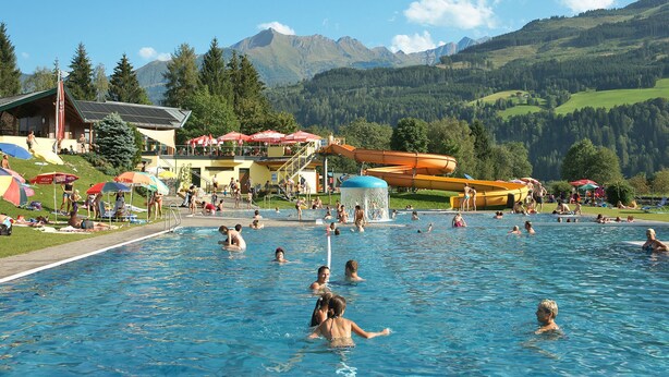 Una piscina con gente disfrutando, un tobogán acuático y montañas al fondo.
