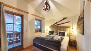 Un dormitorio con una cama grande, vigas de madera, un ventanal con cortinas, una lámpara colgante y un cuadro en la pared.