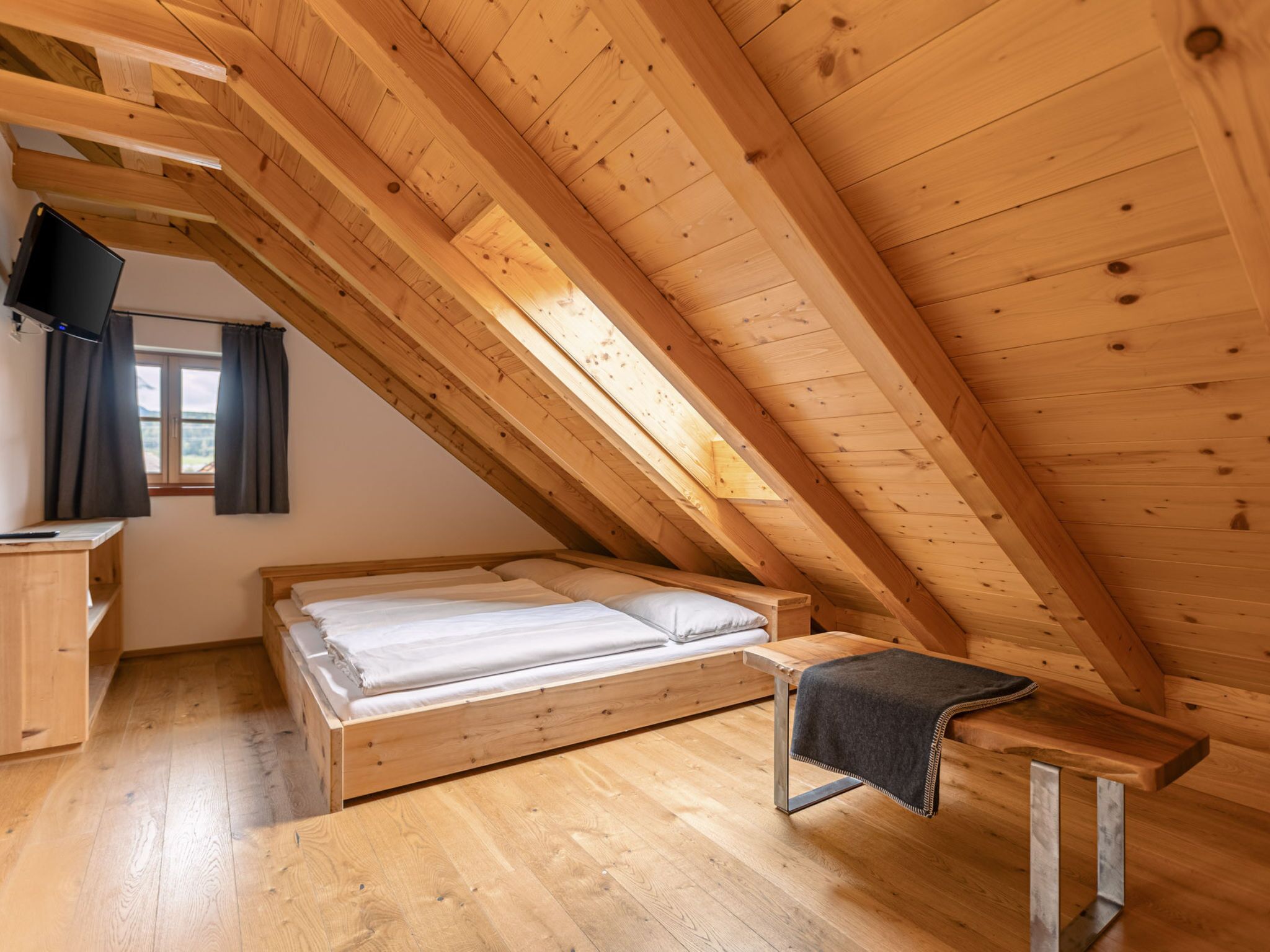 A wooden loft bedroom with a bed, a bench, a TV, and a window.
