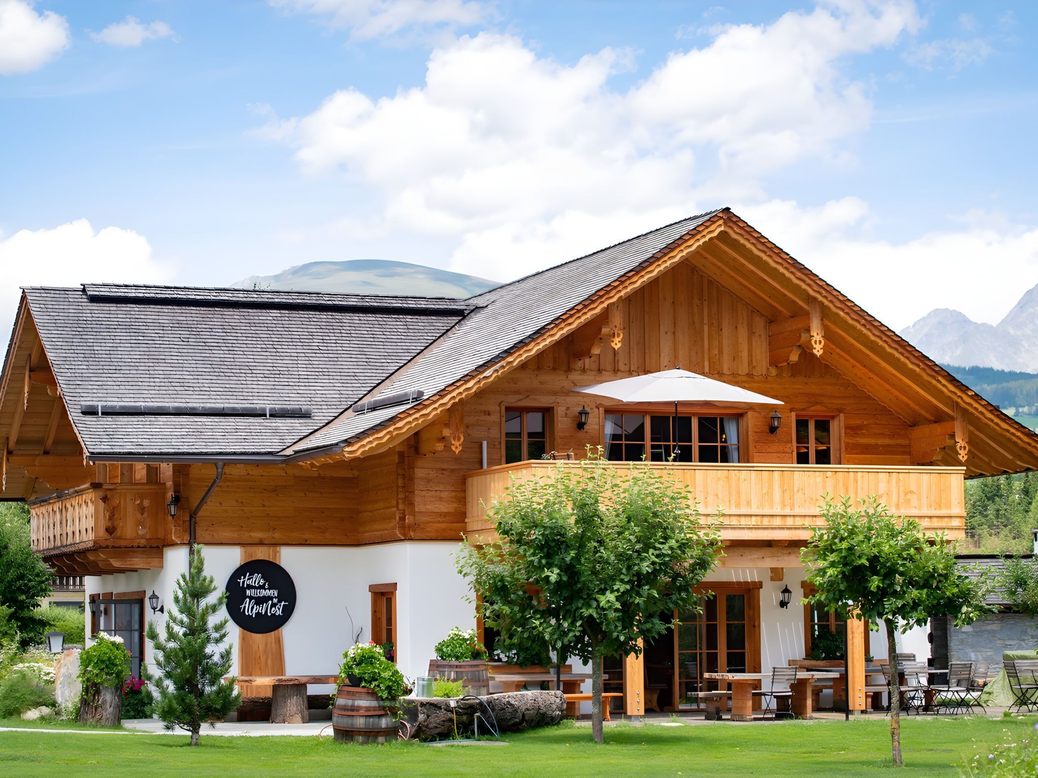 Un chalet en bois doté d’un balcon, une enseigne indiquant « Hall & Nest Alpenhof », et un paysage montagneux en arrière-plan.