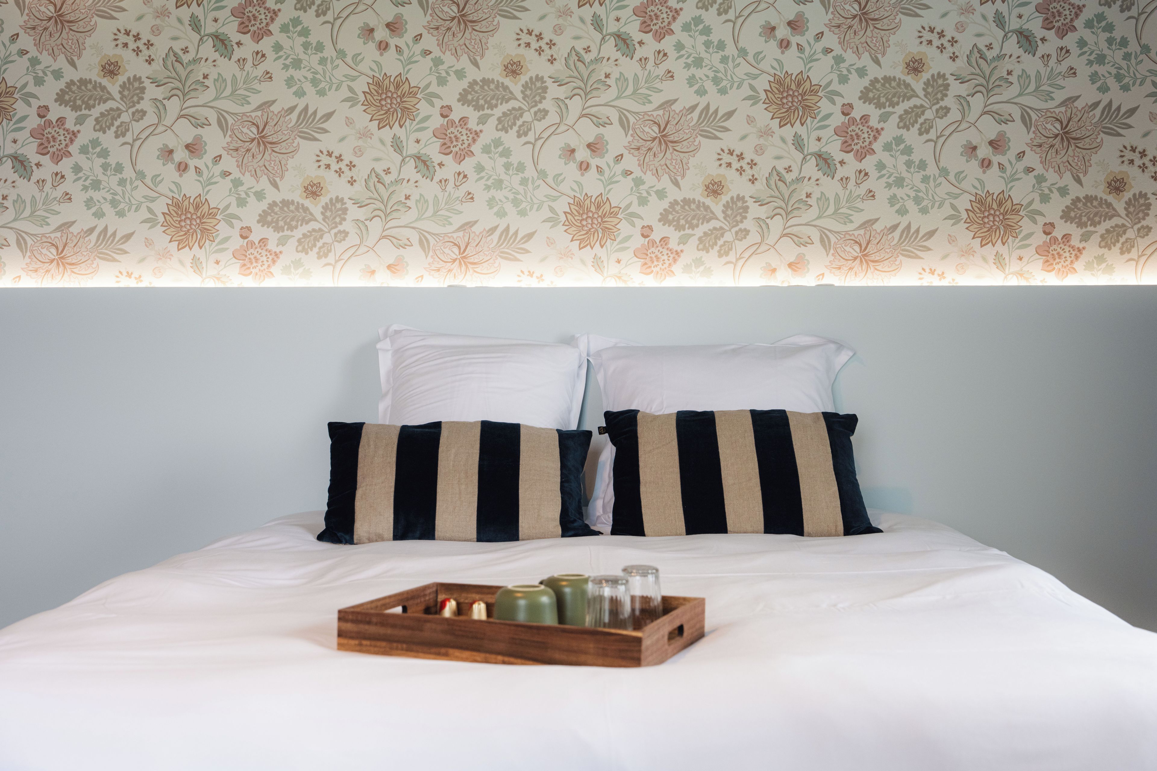 A bed with white bedding and striped pillows, a wooden tray with bottles, and a floral wallpaper pattern.
