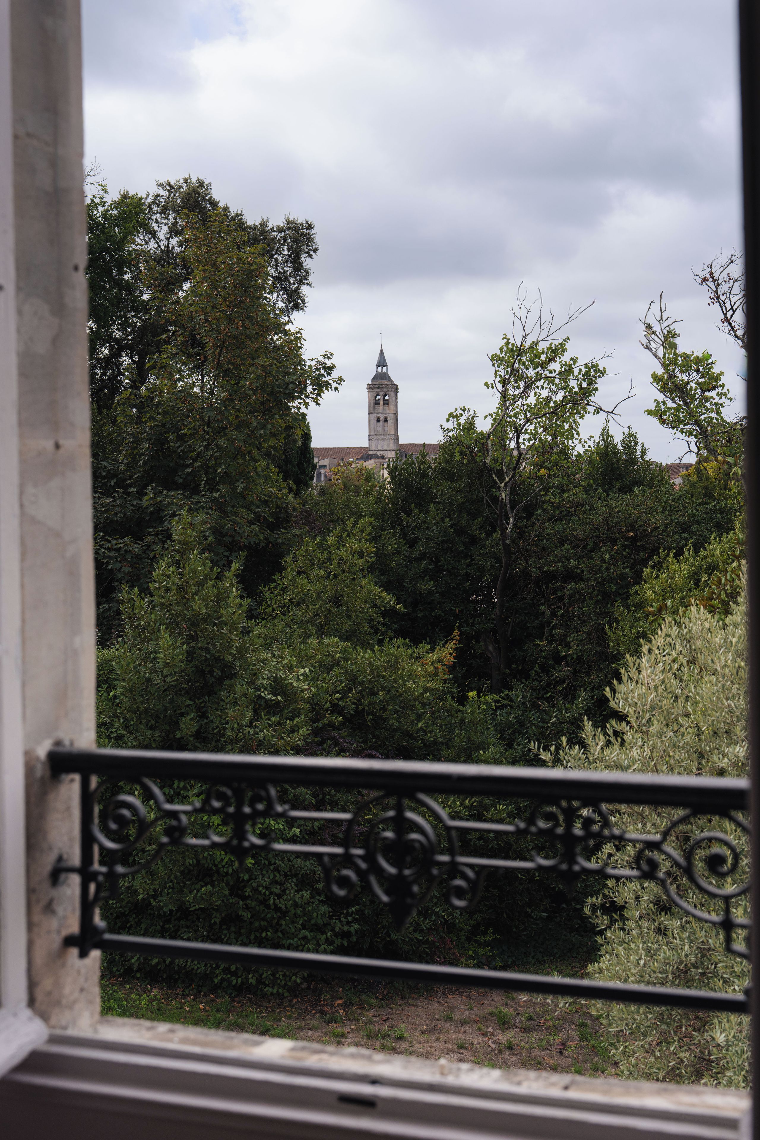 Vista attraverso una finestra con ringhiera in metallo, su un edificio con una torre e vegetazione lussureggiante.