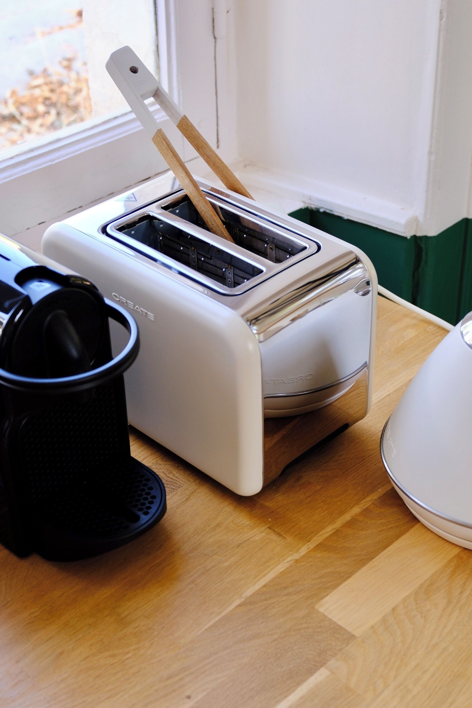 A kitchen with a toaster on a wooden table, a black object on the left, and a window in the background.
