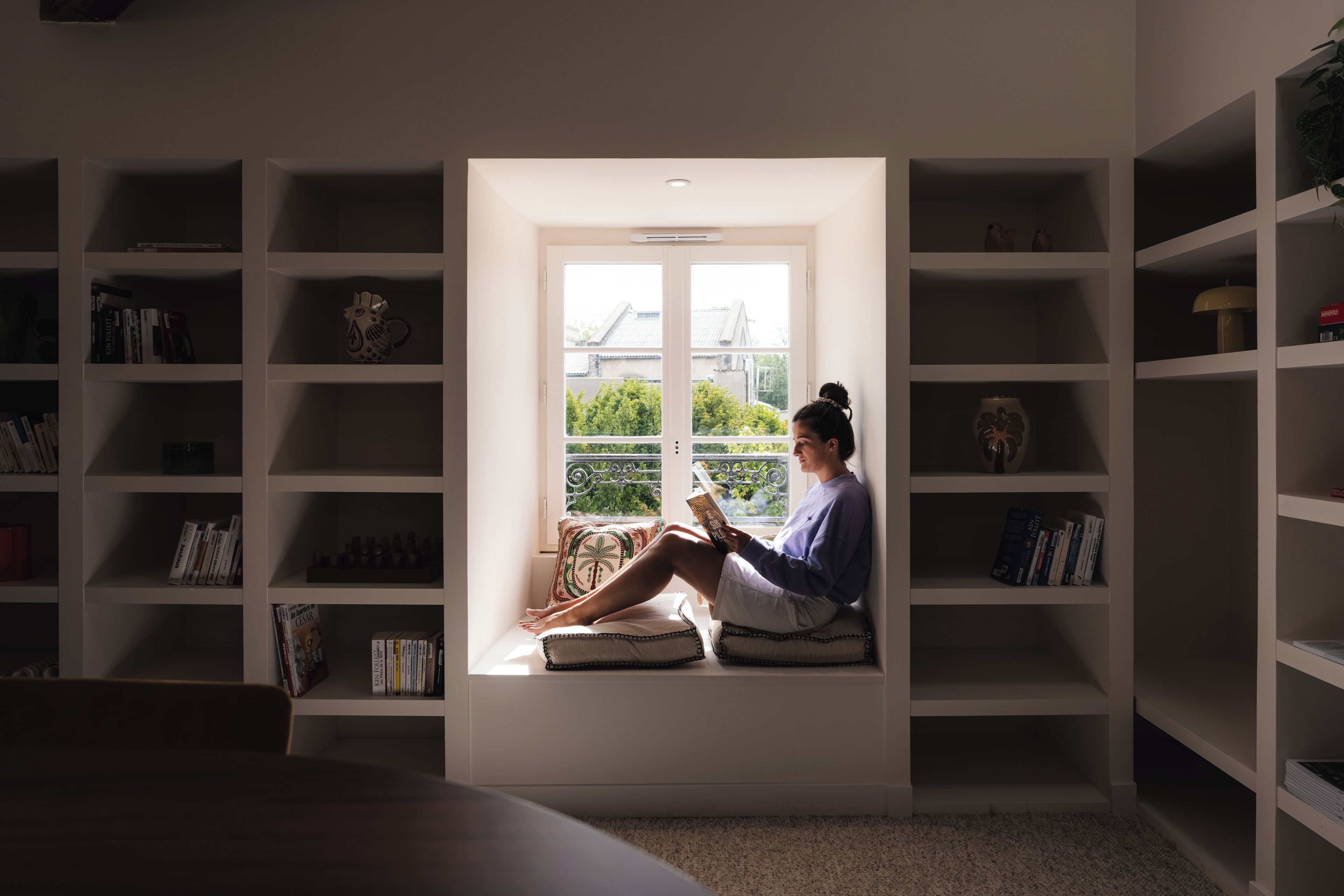Una persona leyendo un libro en una habitación con un ventanal grande, rodeada de estanterías.