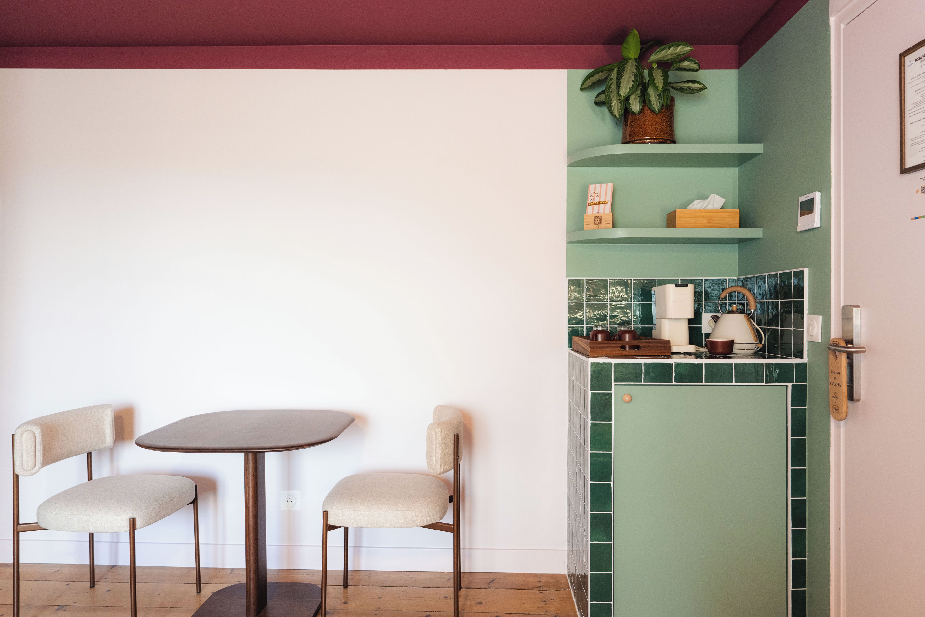 Een kleine keuken met een groene betegelde achterwand, een houten kastje en een aanrecht met een waterkoker en kopjes.