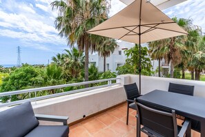 Ein Balkon mit Tisch und Stühlen, einem großen Sonnenschirm und Blick auf Palmen und Gebäude.