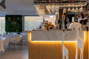 Un'area bar moderna con un bancone in legno, diversi bicchieri e vista su una sala da pranzo con sedie e tavoli bianchi.