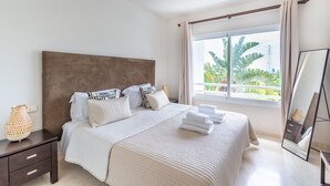 Ein Schlafzimmer mit einem großen Bett, einem Nachttisch, einer Nachttischlampe, einem Fenster mit Vorhängen und einem Spiegel.