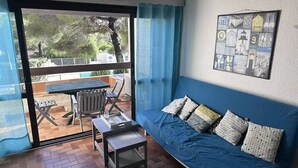 A room with a blue sofa, a small table, and a view through a glass door to an outdoor patio with a table and chairs. - Appartement  Résidence Acapulco avec piscine (Les Issambres)