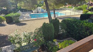 A swimming pool surrounded by a white fence, with a building in the background and various trees and shrubs in the foreground. - Appartement  Résidence Acapulco avec piscine (Les Issambres)