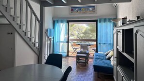A room with a dining table, chairs, a bed, a staircase, and a view through a glass door to an outdoor area with a table and chairs. - Appartement  Résidence Acapulco avec piscine (Les Issambres)
