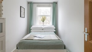 A bedroom with a bed, a window with curtains, a bedside table, a door, and a wall-mounted picture. - Hampstead Heath 2 bed (Greater London)