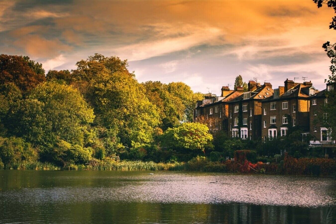 A lakeside view with a row of traditional brick buildings, calm water, and dense greenery.