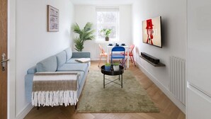 A living room with a sofa, a coffee table, a TV, a plant, and a dining area with chairs. - Hampstead Heath 2 bed (Greater London)