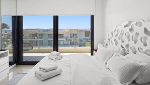 Un dormitorio moderno con una cama grande, vistas de edificios a través de ventanales de techo a suelo y un balcón con una silla.