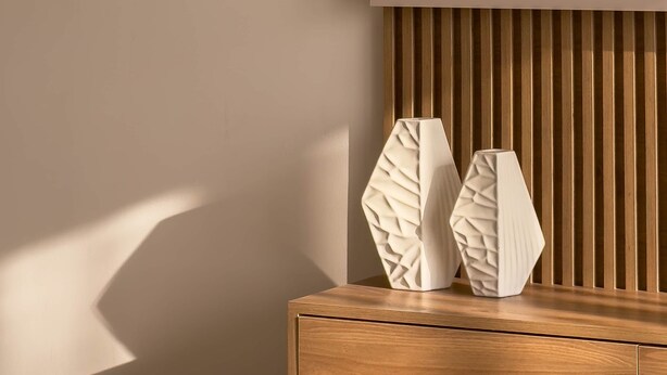 Una mesita de noche de madera con dos jarrones blancos y con textura, y un panel de madera con listones al fondo.