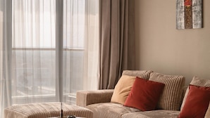 A modern living room with a sofa, ottoman, and coffee table, large windows with sheer curtains, and a piece of abstract art on the wall. - AEON Towers Lucky Cloud #606 - nesty (6th of October City)
