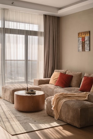 A modern living room with a sofa, ottoman, and coffee table, large windows with sheer curtains, and a piece of abstract art on the wall.