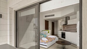 A modern hotel room with a sliding glass door leading to a balcony, a small round table, a bed with colorful bedding, and a kitchenette area.