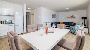 Et moderne køkken med hvide skabe, en mikrobølgeovn og et køleskab. Der er et spisebord med fire stole. En sofa og en mønstret skammel ses i baggrunden.