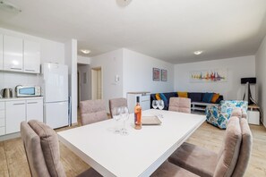 A modern kitchen with white cabinets, a microwave, and a refrigerator. There is a dining area with a white table and four chairs. A sofa and a patterned ottoman are in the background. - 4-star penthouse apartment near the beach (Dramalj)