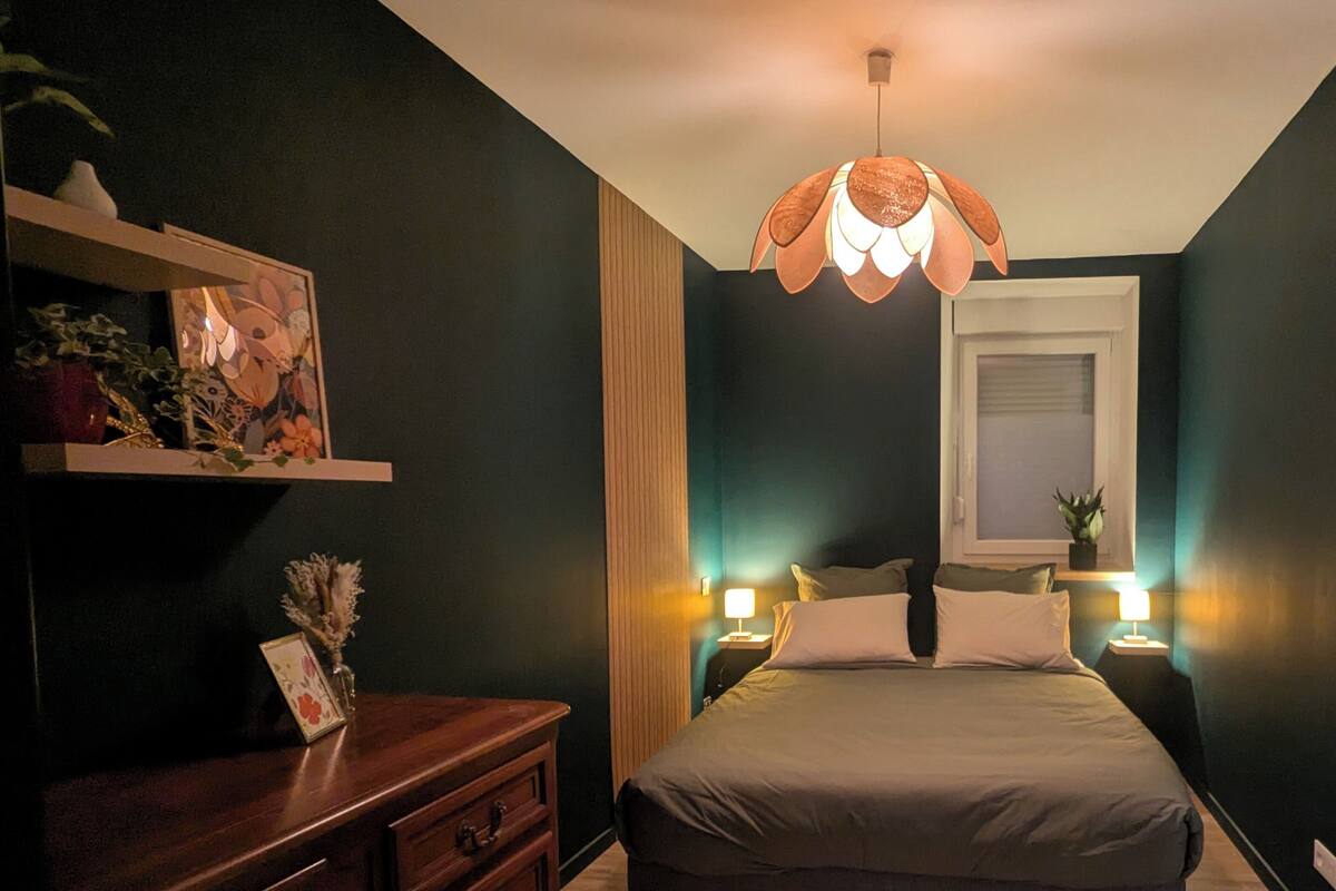 A bedroom with a bed, bedside lamps, a dresser, a hanging light fixture, and a window with blinds.