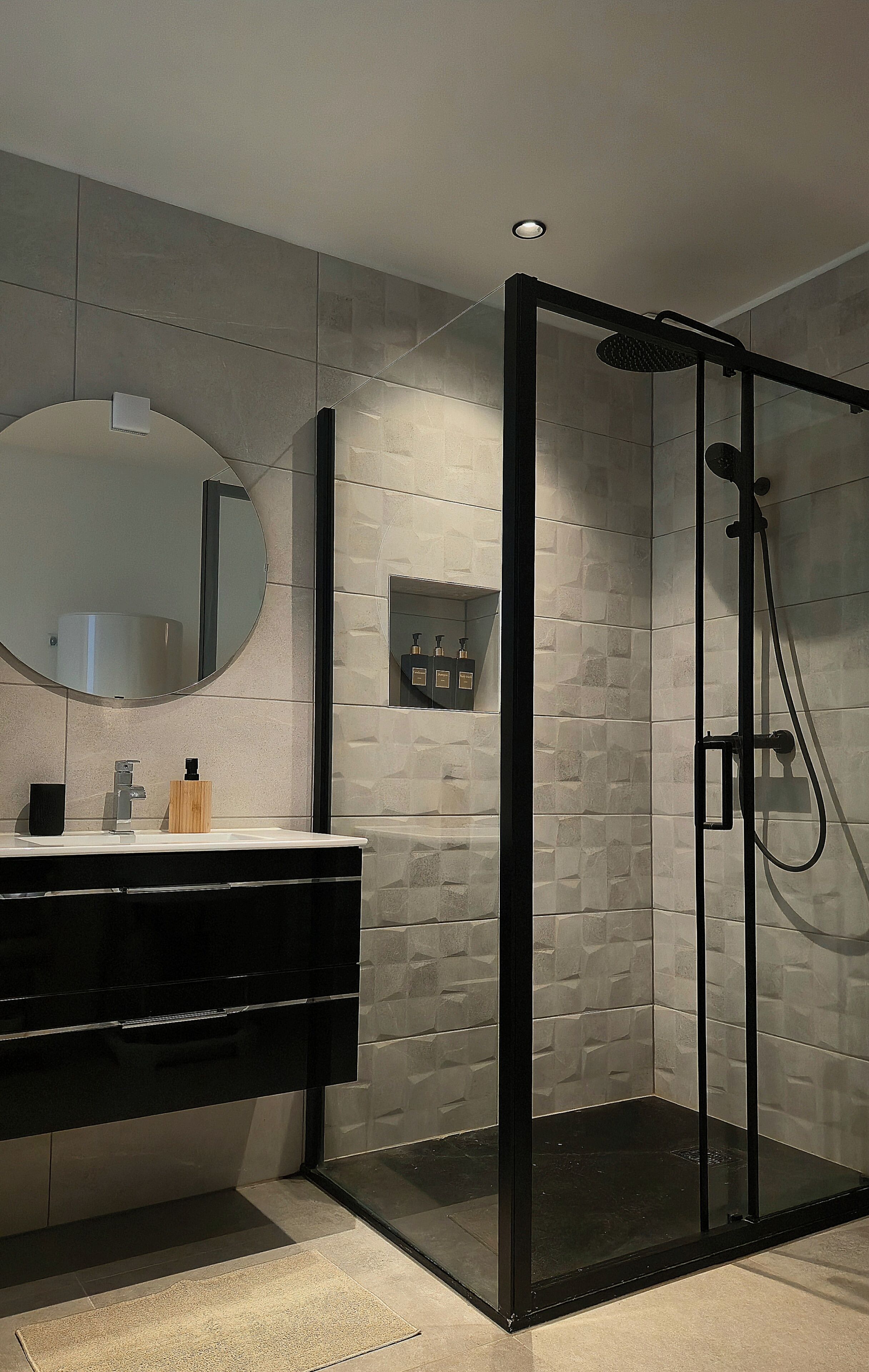 Un baño moderno con ducha de vidrio, espejo redondo y tocador con encimera negra.