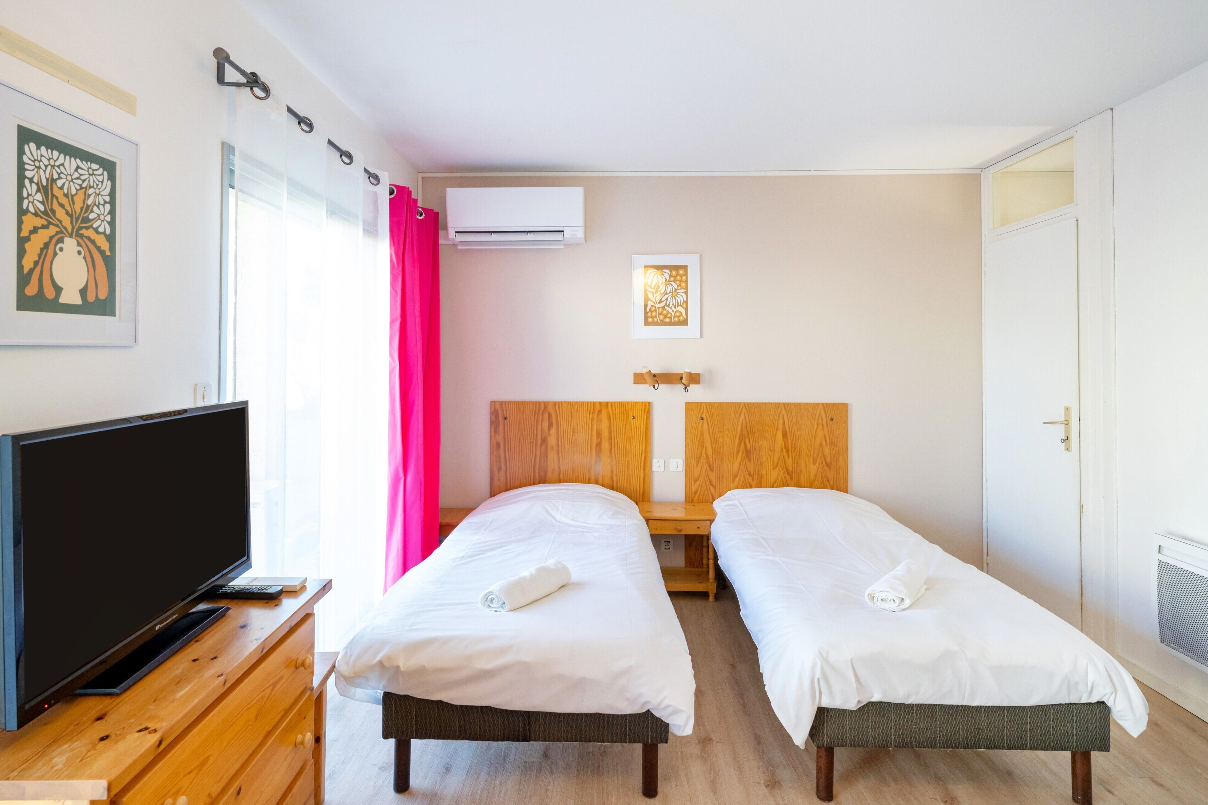 Une chambre d’hôtel avec deux lits simples, une télévision, une commode en bois, un appareil de climatisation et une fenêtre avec un rideau.