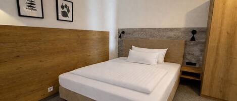 A bedroom with a wooden headboard, a bed with white bedding, two framed pictures on the wall, and a small bedside table.
