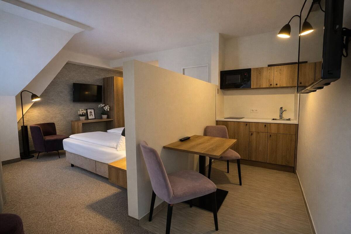 A compact hotel room with a bed, a desk, a chair, a kitchenette, and a TV mounted on the wall.