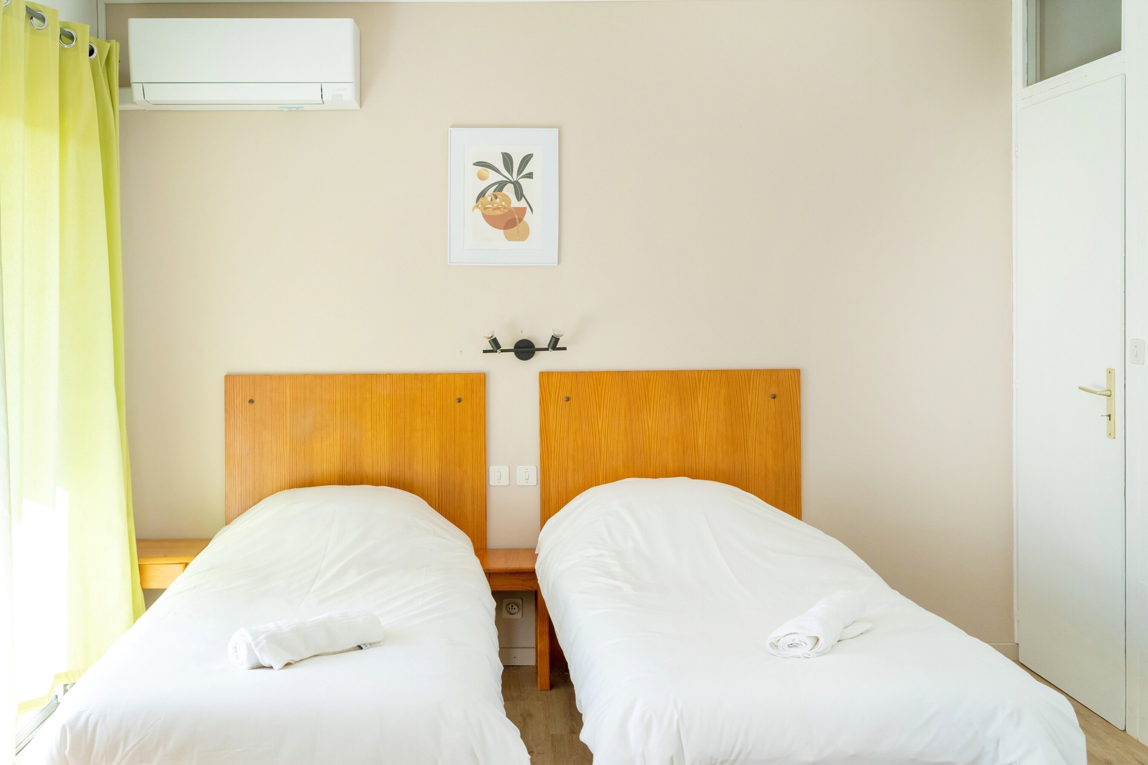 A hotel room with two single beds, a wooden headboard, a small bedside table, a wall-mounted air conditioner, and a framed picture.