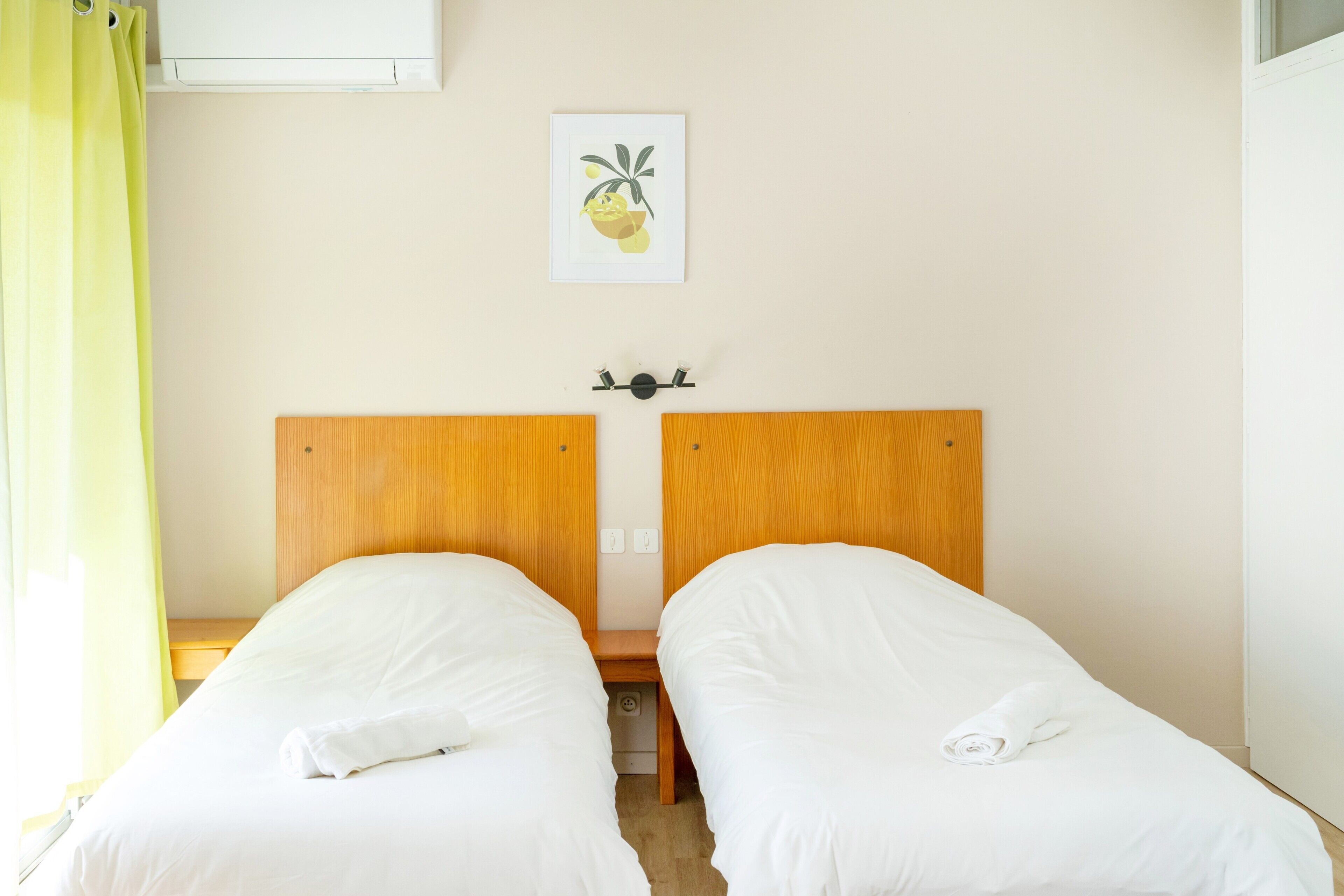 Une chambre avec deux lits simples, une tête de lit en bois, une petite table de chevet, un climatiseur fixé au mur et un tableau encadré.
