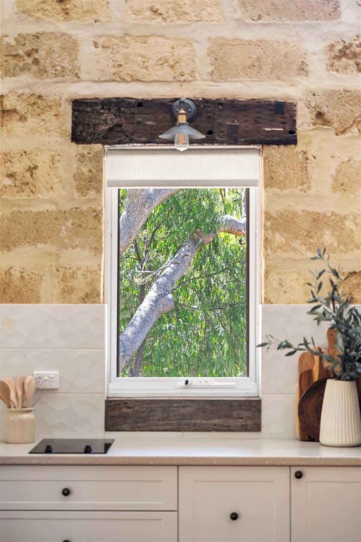 A kitchen with a stone wall, a window with a view of trees, a wooden beam above the window, and a potted plant on the counter.