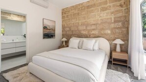 Une chambre avec un mur en briques, un lit, des tables de chevet avec des lampes, un tableau, et une salle de bain avec un miroir et un lavabo.